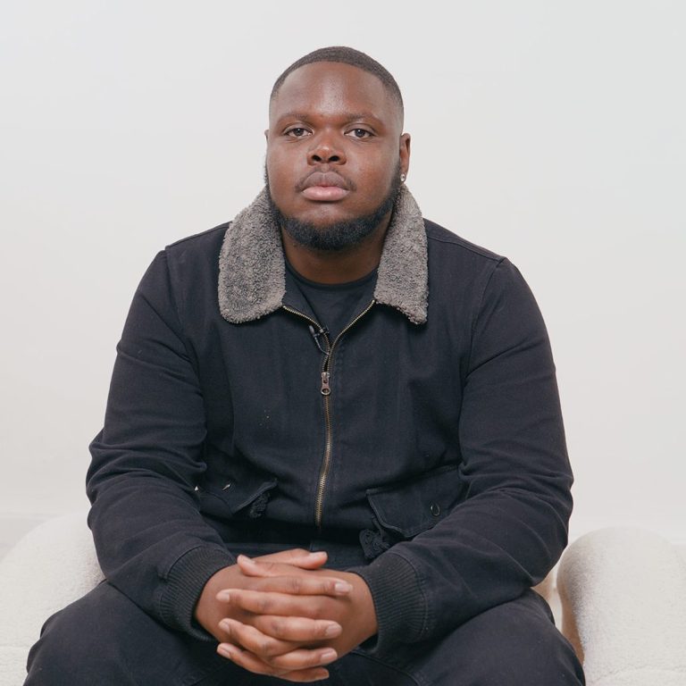 A seated man, wearing a black jacket with a fur collar.