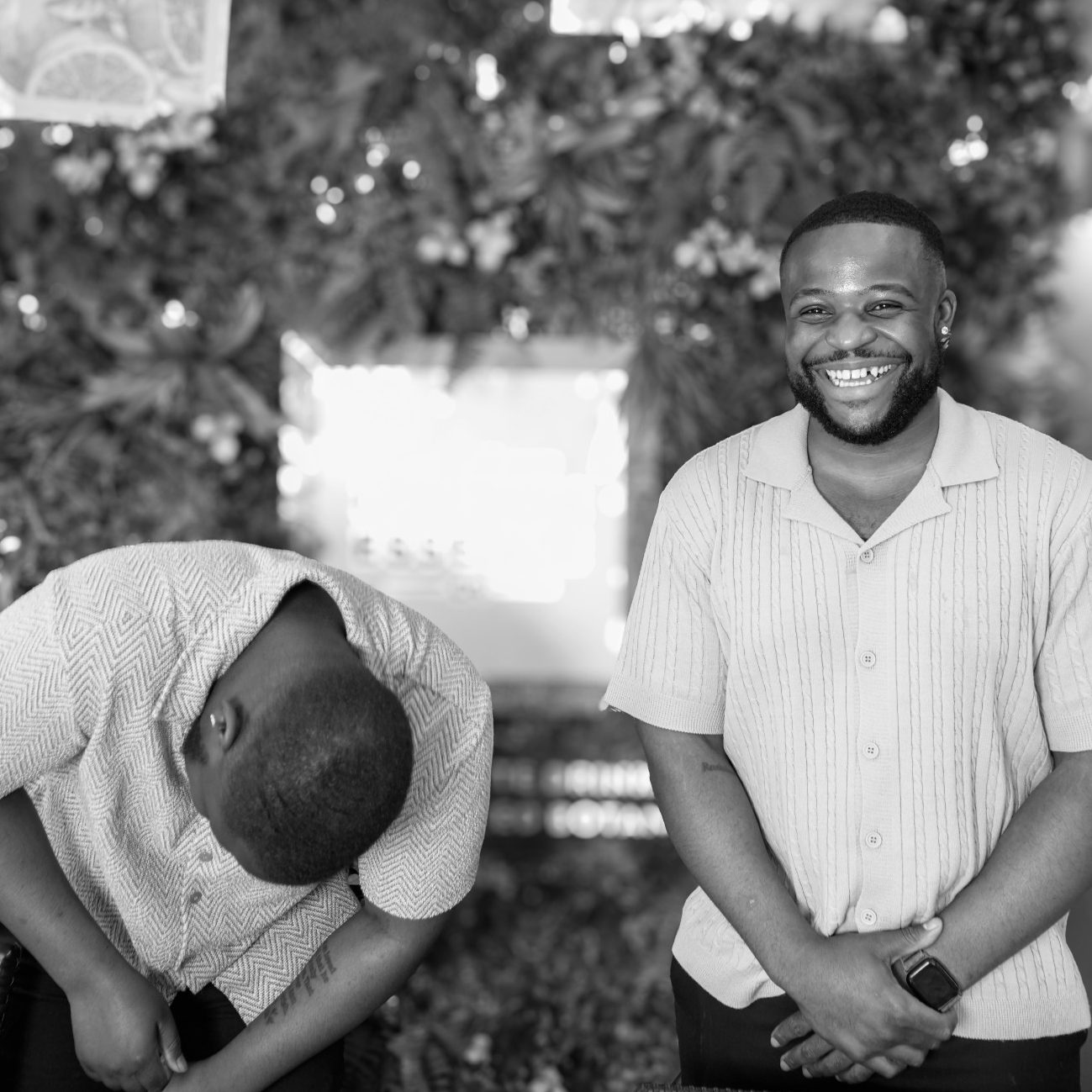 Two men smiling, one leaning forward and the other standing upright, in a floral setting.