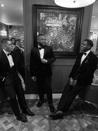 Three men in formal black tuxedos, chatting in an elegant indoor setting.