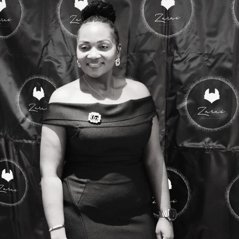 A woman in an elegant black off-shoulder dress smiling in front of a patterned backdrop.