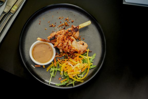 Grilled chicken skewer with sauce, salad, and garnishes on a black plate.