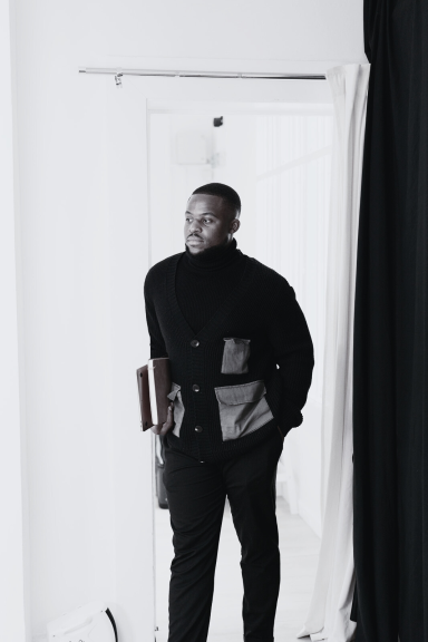 A man in a black coat stands confidently in a stylish indoor setting.